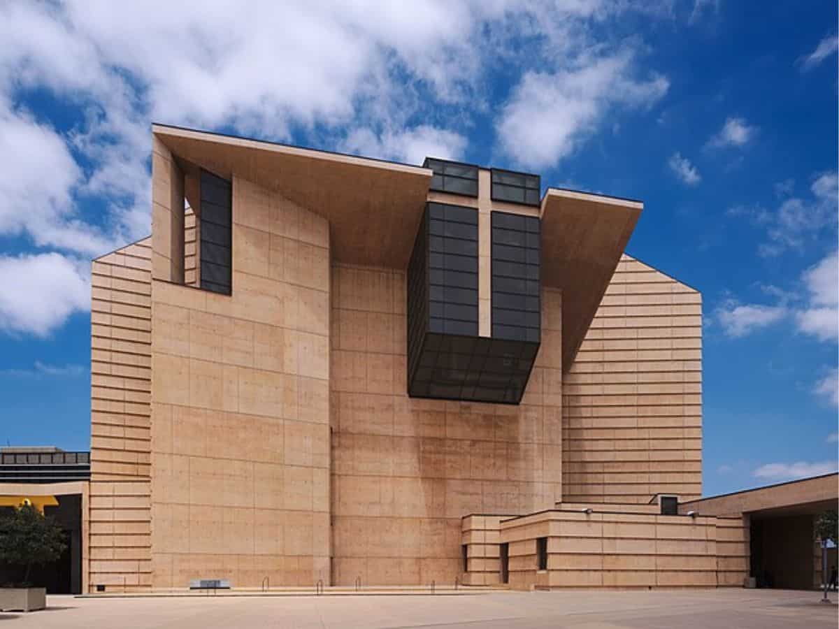 The grand facade of a modern cathedral with angular architectural design and a large cross integrated into the structure, set against a bright blue sky.