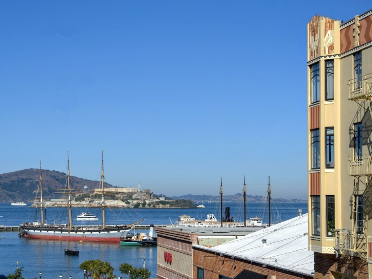 A scenic view of the San Francisco Bay featuring historic sailing ships docked near the waterfront and Alcatraz Island in the background, with a vintage-style building in the foreground. The serene ocean setting and iconic landmarks exemplify the city's premium coastal real estate, raising the question, Why Is San Francisco So Expensive, as the desirable waterfront views significantly contribute to high property values.