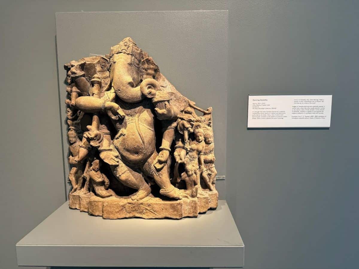 A detailed stone sculpture of Dancing Ganesha displayed in a museum, with an informational plaque nearby. The intricate craftsmanship reflects the cultural richness and diversity of San Francisco's museums and galleries, contributing to the city's vibrant arts scene and raising the question, Why Is San Francisco So Expensive, by showcasing the value placed on maintaining world-class cultural and entertainment attractions.