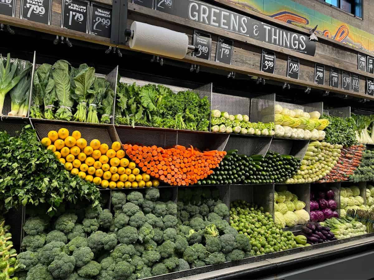 A colorful and neatly arranged produce section in a grocery store labeled "Greens & Things," showcasing fresh vegetables like broccoli, carrots, zucchini, and leafy greens alongside vibrant fruits. The abundance of high-quality, organic produce reflects San Francisco's emphasis on premium food options, contributing to the question, Why Is San Francisco So Expensive, as seen in the elevated cost of groceries in the area.