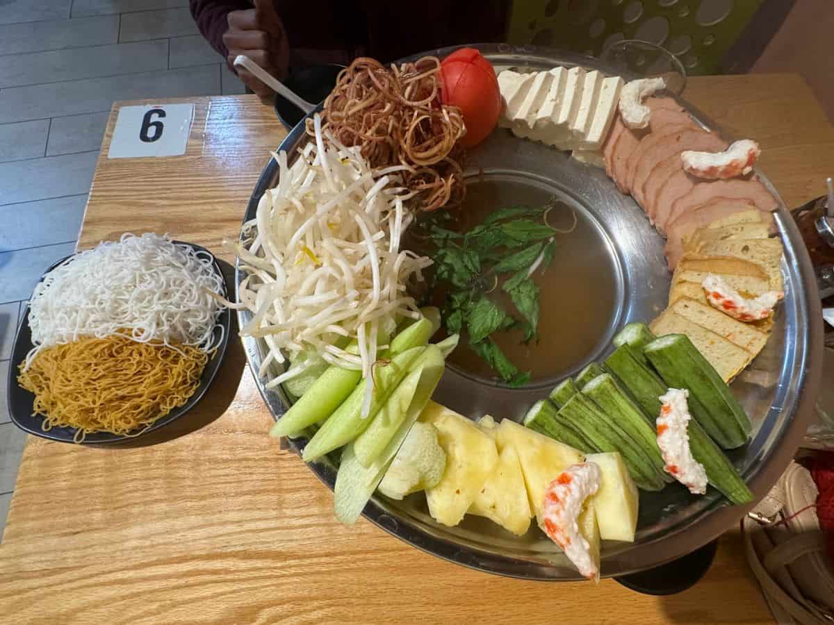 A colorful spread of fresh vegan ingredients arranged around a hotpot at one of the 10 Vegan Spots in San Jose. The platter includes bean sprouts, okra, cucumber, pineapple, fresh tofu slices, vegan ham, and tofu-based seafood alternatives, accompanied by fresh herbs. On the side, a plate features a variety of noodles, including white rice noodles and yellow eggless noodles, ready to be cooked in the savory broth at the center.