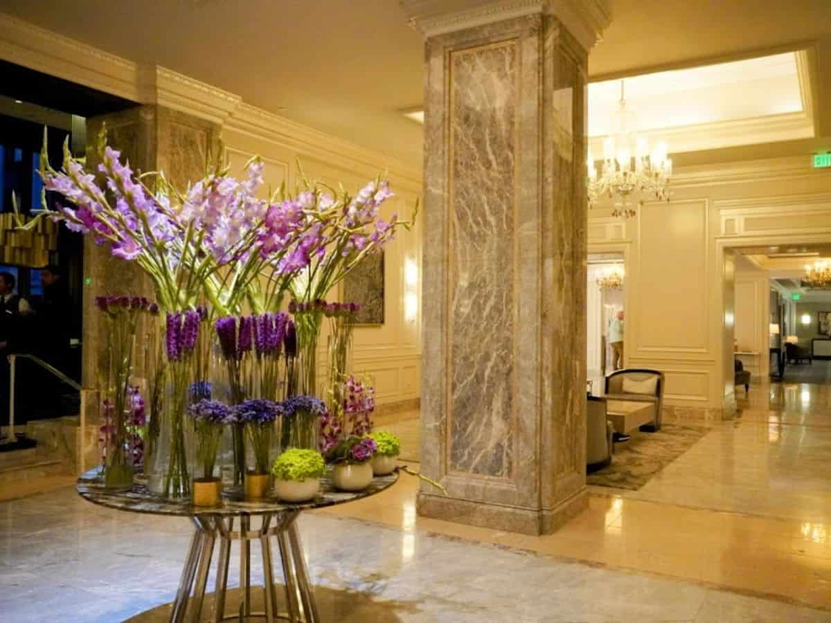 A luxurious hotel lobby with polished marble floors, grand pillars, and an elegant floral arrangement of vibrant purple and green blooms displayed on a central table. The opulent decor reflects the upscale business and hospitality sector in San Francisco, raising the question, Why Is San Francisco So Expensive, as high business taxes and premium service expectations drive up operating costs that influence prices citywide.