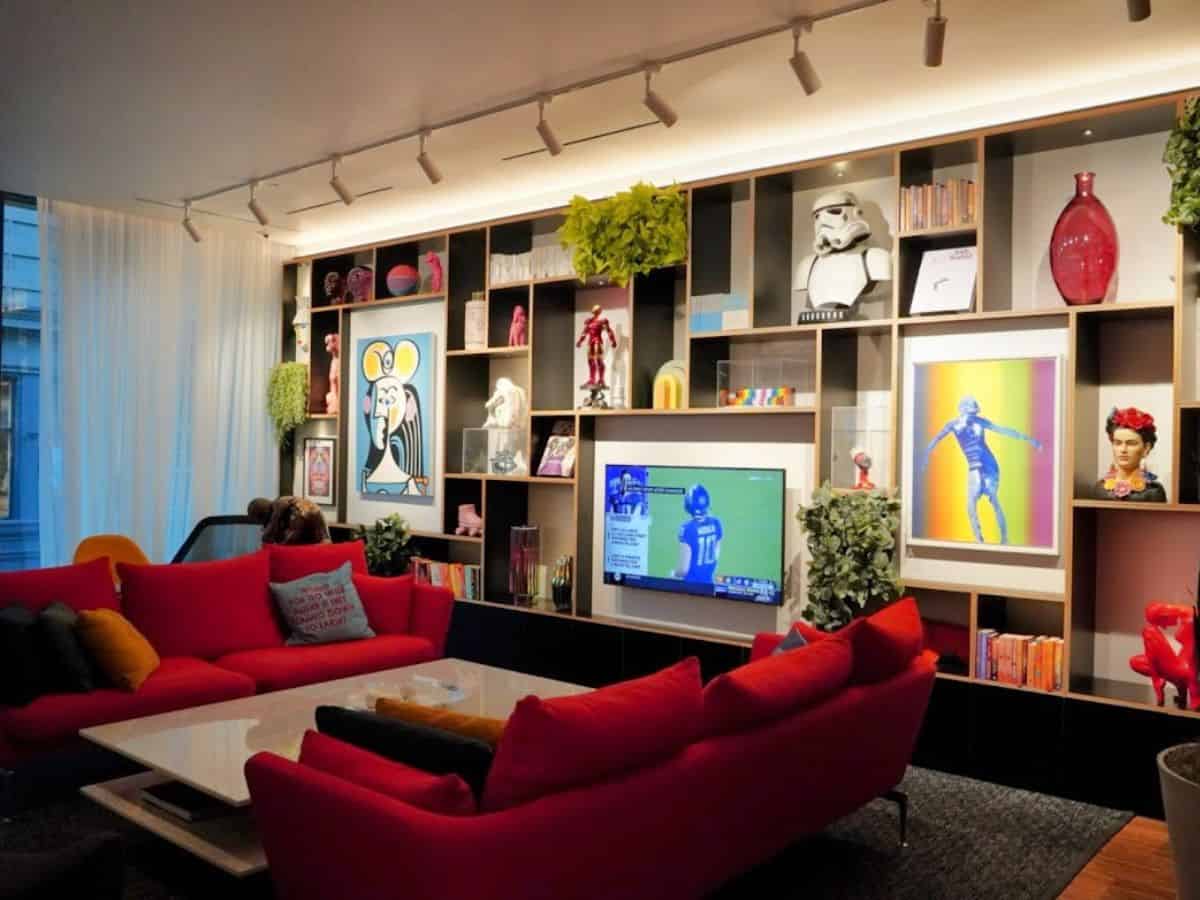 A modern, stylish living room with vibrant red sofas, artistic decor, and a wall-mounted TV surrounded by a display of eclectic books, plants, and collectibles. The luxurious and trendy interior exemplifies the upscale living spaces in San Francisco, emphasizing the high demand for premium housing and sparking the question, Why Is San Francisco So Expensive, due to the city's focus on aesthetics and lifestyle.