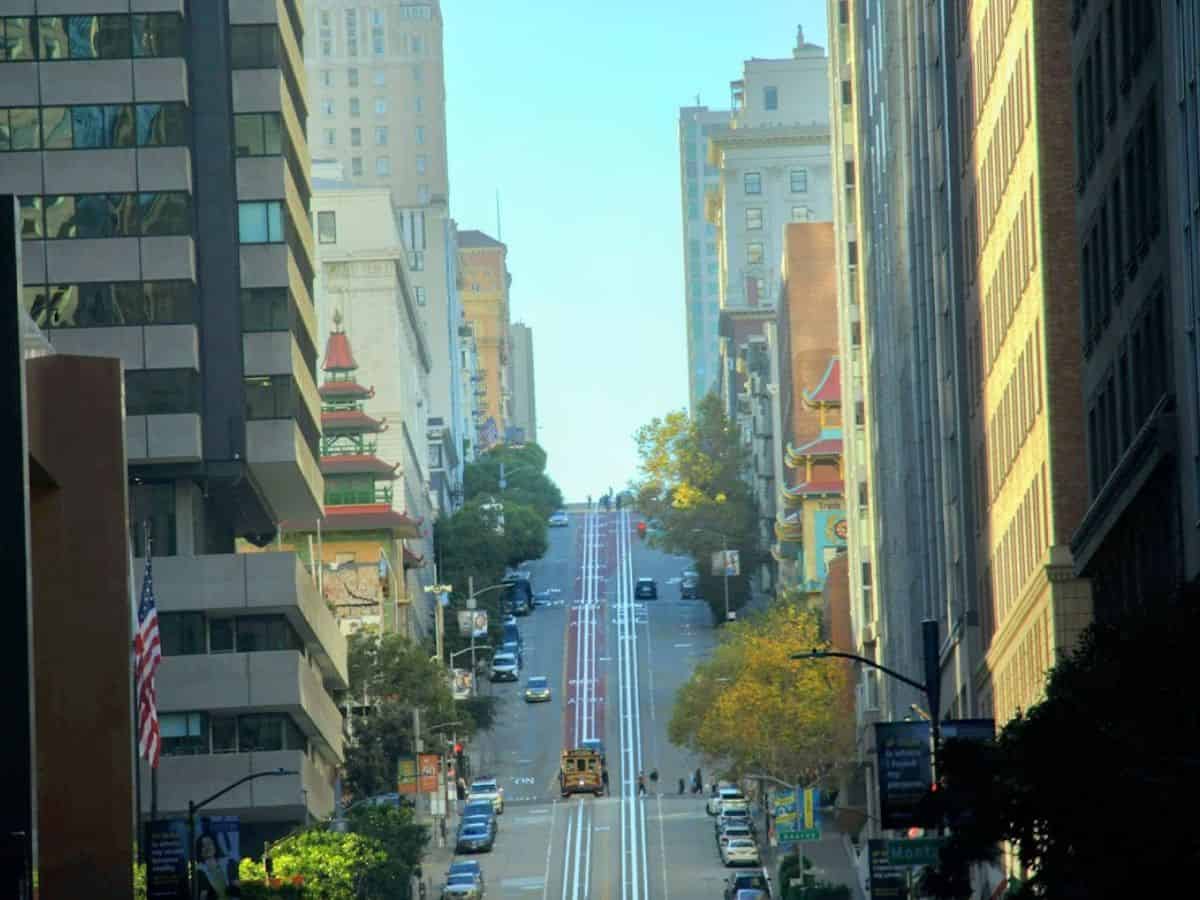 A picturesque view of a steep San Francisco street lined with tall buildings and a cable car making its way up the hill. The unique architecture and iconic slopes, with a touch of Chinatown's pagoda-style elements, highlight the city's distinctive geography and urban charm, raising the question, Why Is San Francisco So Expensive, as the terrain and infrastructure demands contribute to higher living costs.