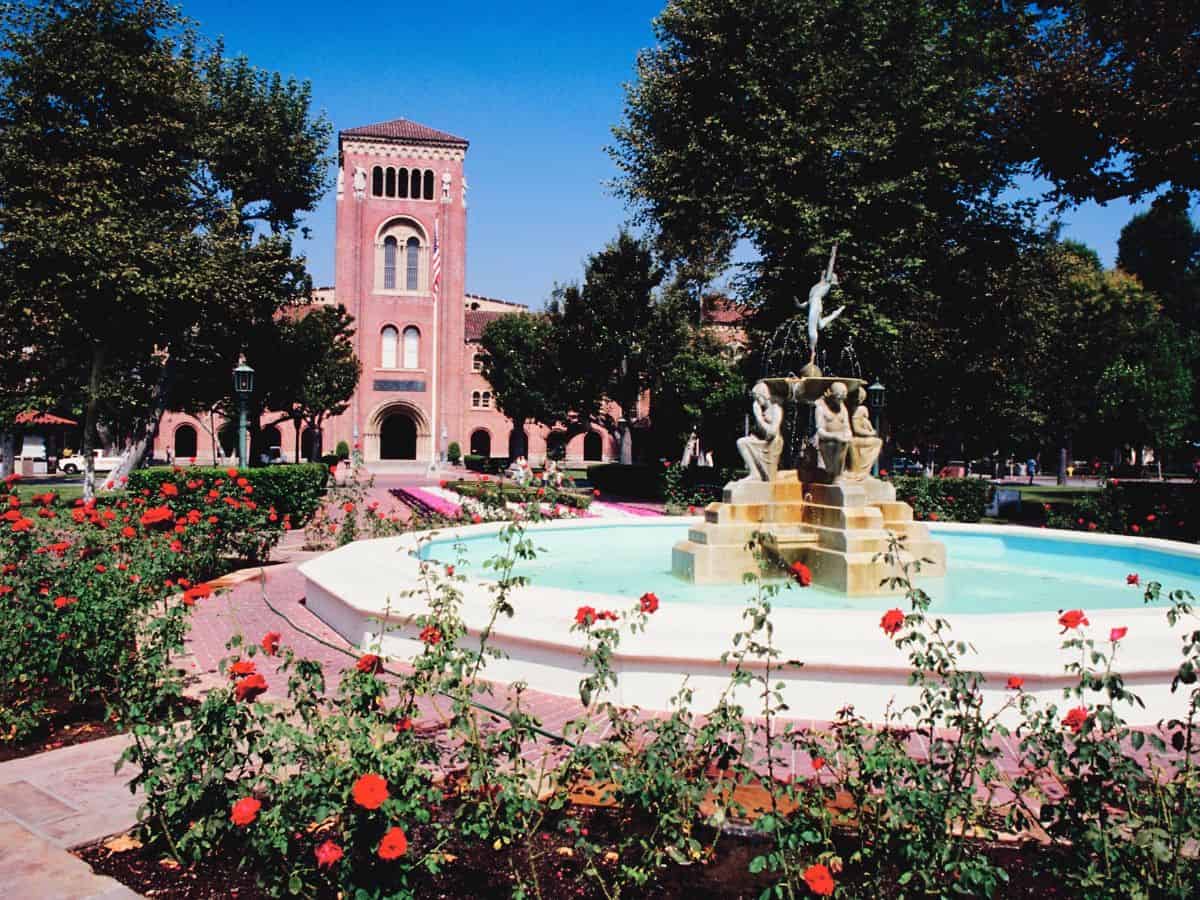 A picturesque view of a university campus in Los Angeles, featuring a historic brick building, a fountain adorned with sculptures, and vibrant rose gardens. The serene yet prestigious setting highlights the allure of higher education in the city while drawing attention to its financial demands, contributing to the question: Why Is Los Angeles So Expensive? The Truth About the High Costs of Living.