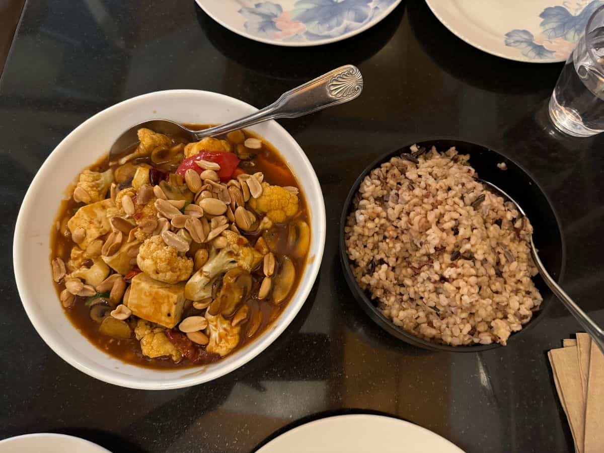A flavorful vegan meal from one of the 10 Vegan Spots in San Jose. The dish features a bowl of rich and savory stew made with cauliflower, tofu, mushrooms, cherry tomatoes, and a sprinkle of roasted peanuts for added texture and crunch. It is paired with a side of hearty multigrain rice, creating a wholesome and satisfying combination.