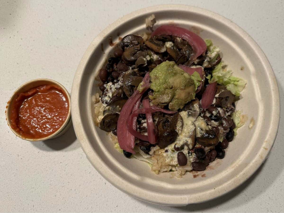 A hearty vegan burrito bowl featuring brown rice, black beans, sautéed mushrooms, pickled red onions, creamy guacamole, and fresh lettuce, topped with a drizzle of vegan cheese sauce. A side of vibrant red salsa adds a zesty touch to this dish, likely served at one of the 10 Vegan Spots in San Jose.