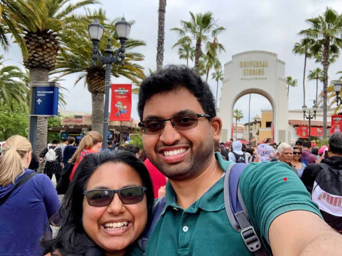 A cheerful couple takes a selfie in front of the iconic entrance arch of Universal Studios Hollywood in Los Angeles, surrounded by a bustling crowd and palm trees. The photo reflects the vibrant atmosphere of a popular tourist destination, highlighting one of the lifestyle perks contributing to the question: Why Is Los Angeles So Expensive? The Truth About the High Costs of Living.