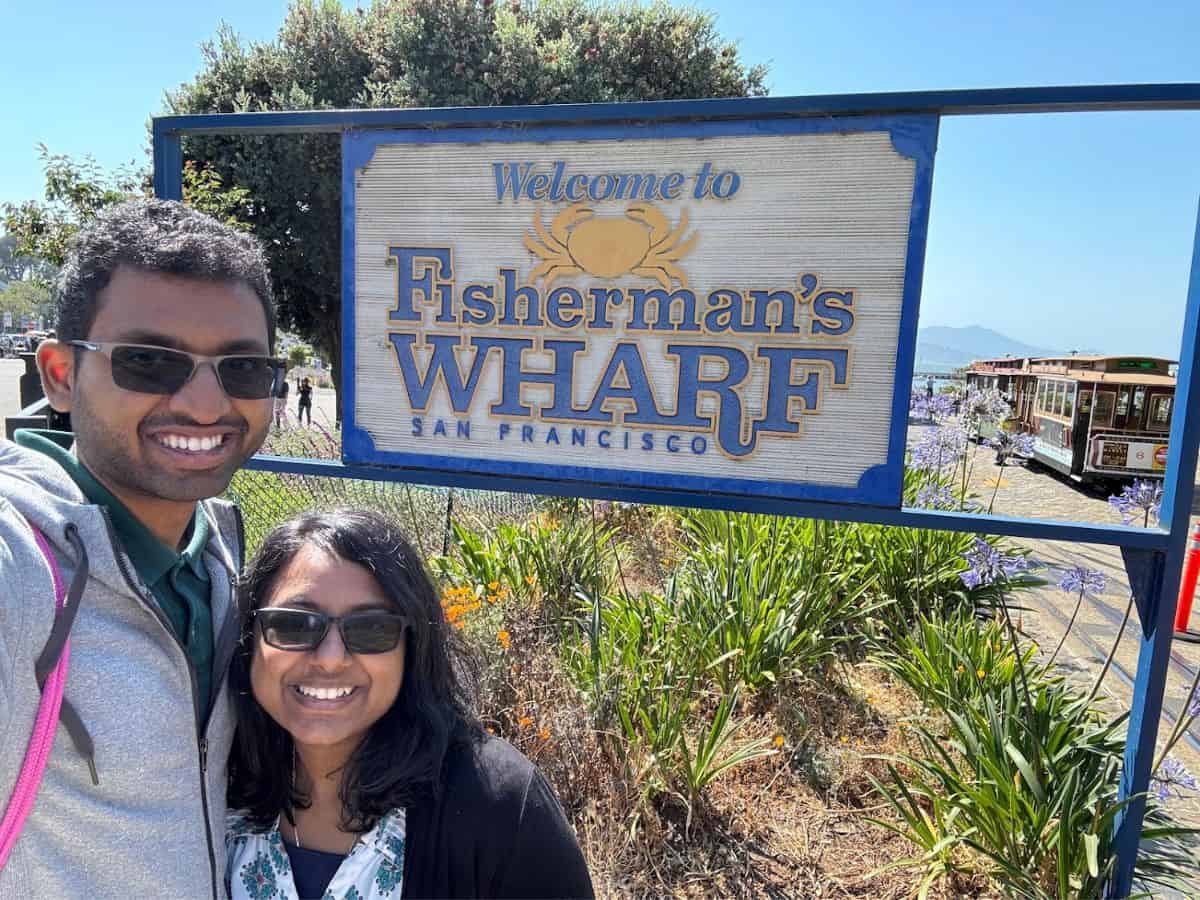 A cheerful couple poses in front of a "Welcome to Fisherman's Wharf, San Francisco" sign on a sunny day. The background features vibrant greenery, iconic cable cars, and a glimpse of the San Francisco Bay, highlighting the city's popular tourist attractions and the question, Why Is San Francisco So Expensive, through its bustling and scenic environment.