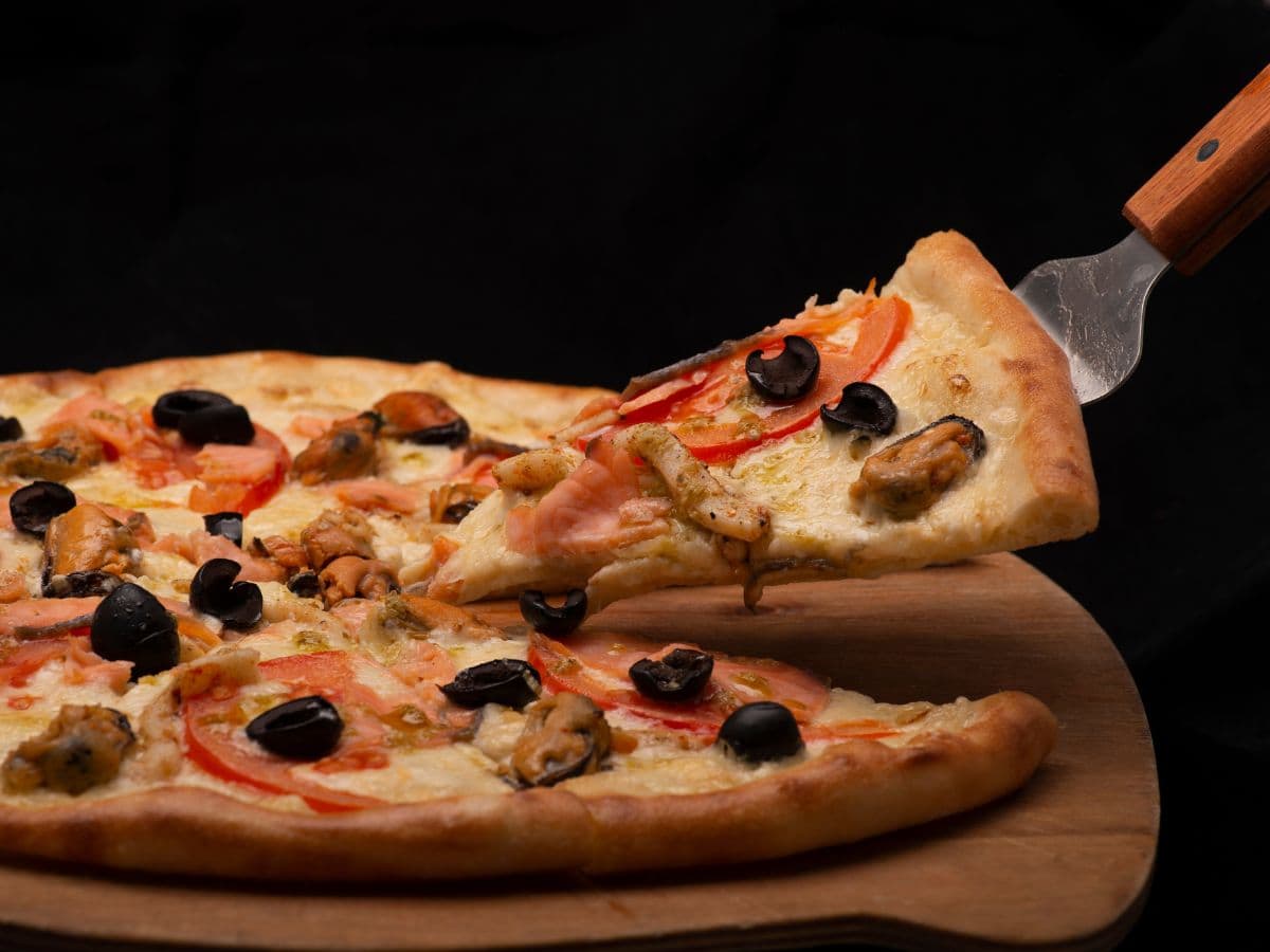 A seafood pizza on a wooden board, topped with shrimp, mussels, sliced tomatoes, and black olives.