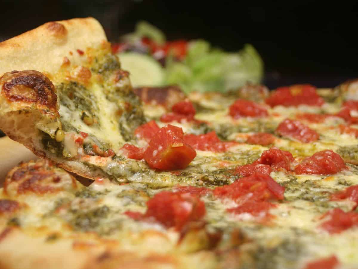 A slice of pesto pizza being lifted, topped with vibrant tomatoes, melted cheese, and green pesto sauce.