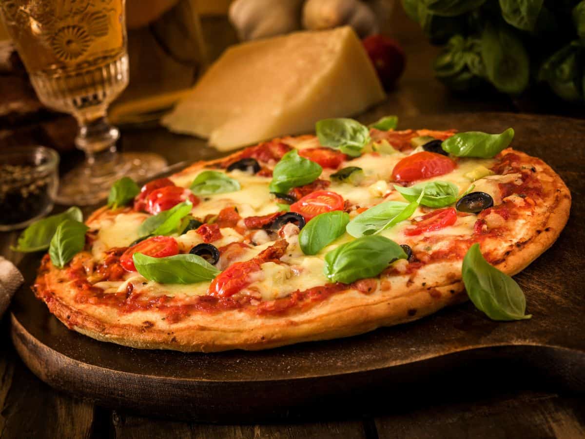 A pizza topped with fresh basil leaves, cherry tomatoes, and black olives, served on a wooden board with cheese and wine in the background.