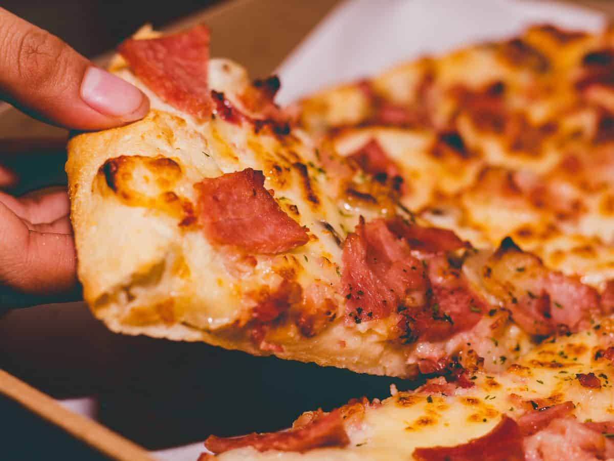 A close-up of a slice of pizza with crispy crust, melted cheese, and bacon, being held in a person's hand.