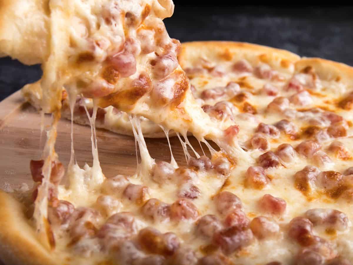 A pizza with crispy crust and bubbling cheese, topped with bacon bits, with a slice being lifted to show the stretchy melted cheese.