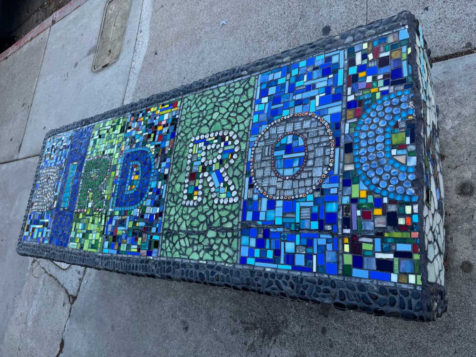 A colorful mosaic bench decorated with intricate glass tiles and patterns, spelling out "BIRD ROCK" in vibrant blues and greens.