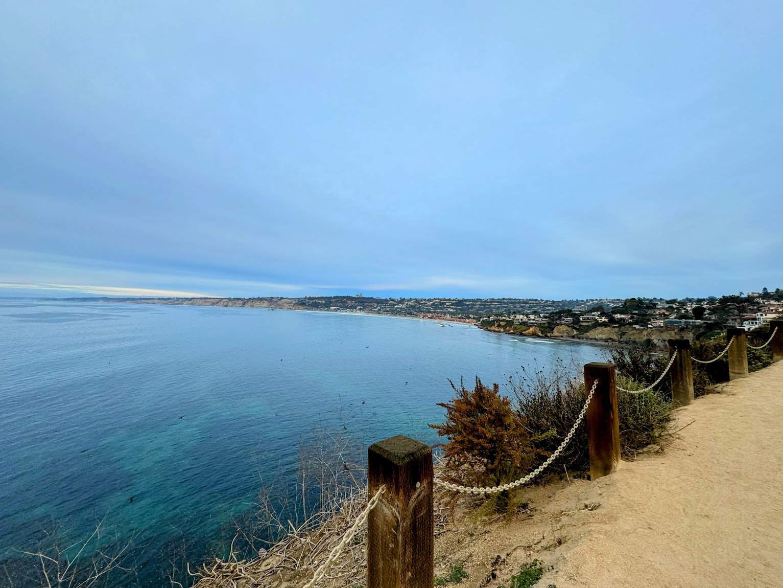 A scenic coastal trail in La Jolla with a dirt path bordered by a rope fence, overlooking calm, blue ocean waters and distant cliffs under a slightly overcast sky.