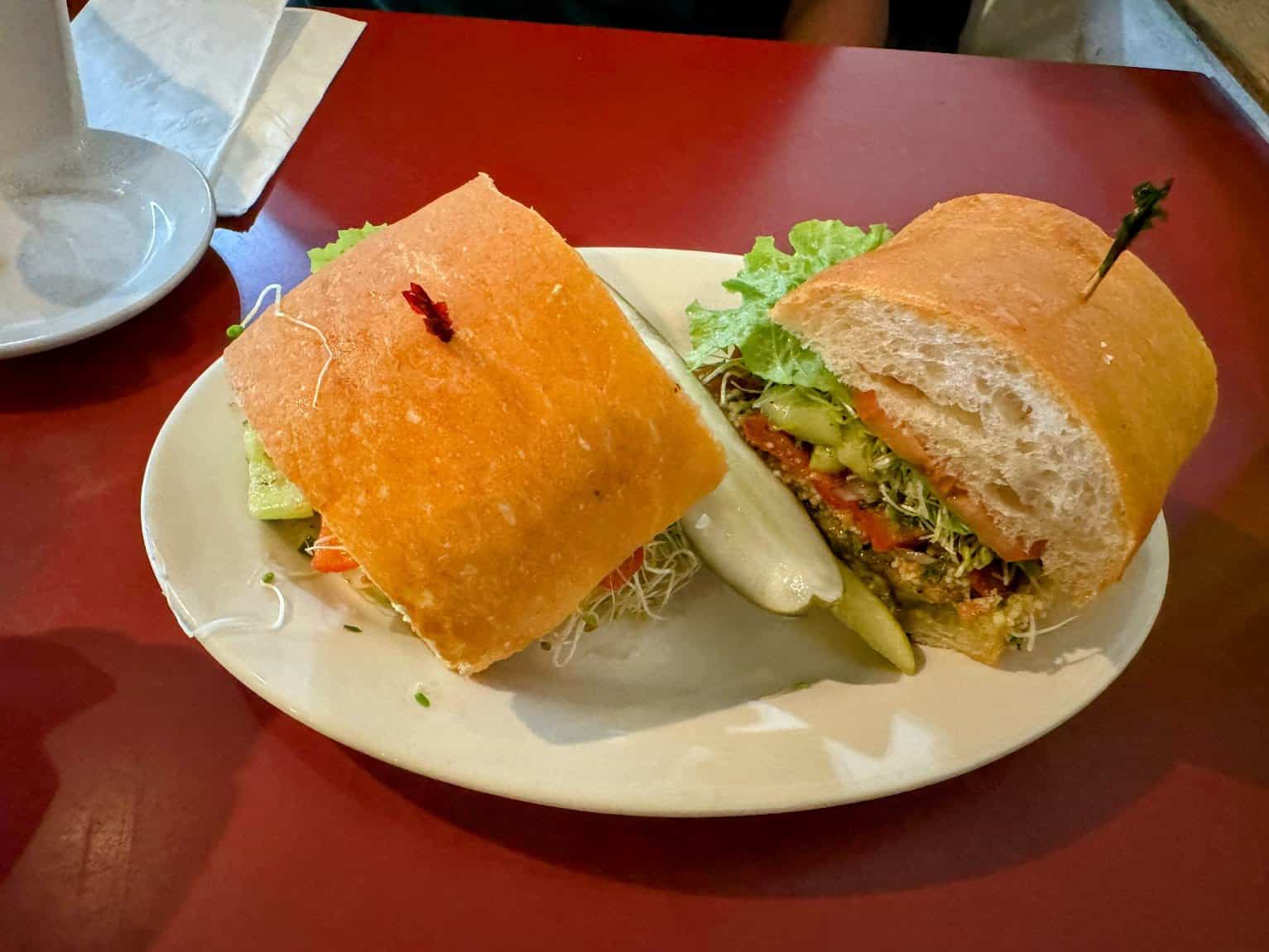 A hearty sandwich with fresh vegetables, sprouts, and lettuce on thick bread, served with a pickle on the side on a white plate.