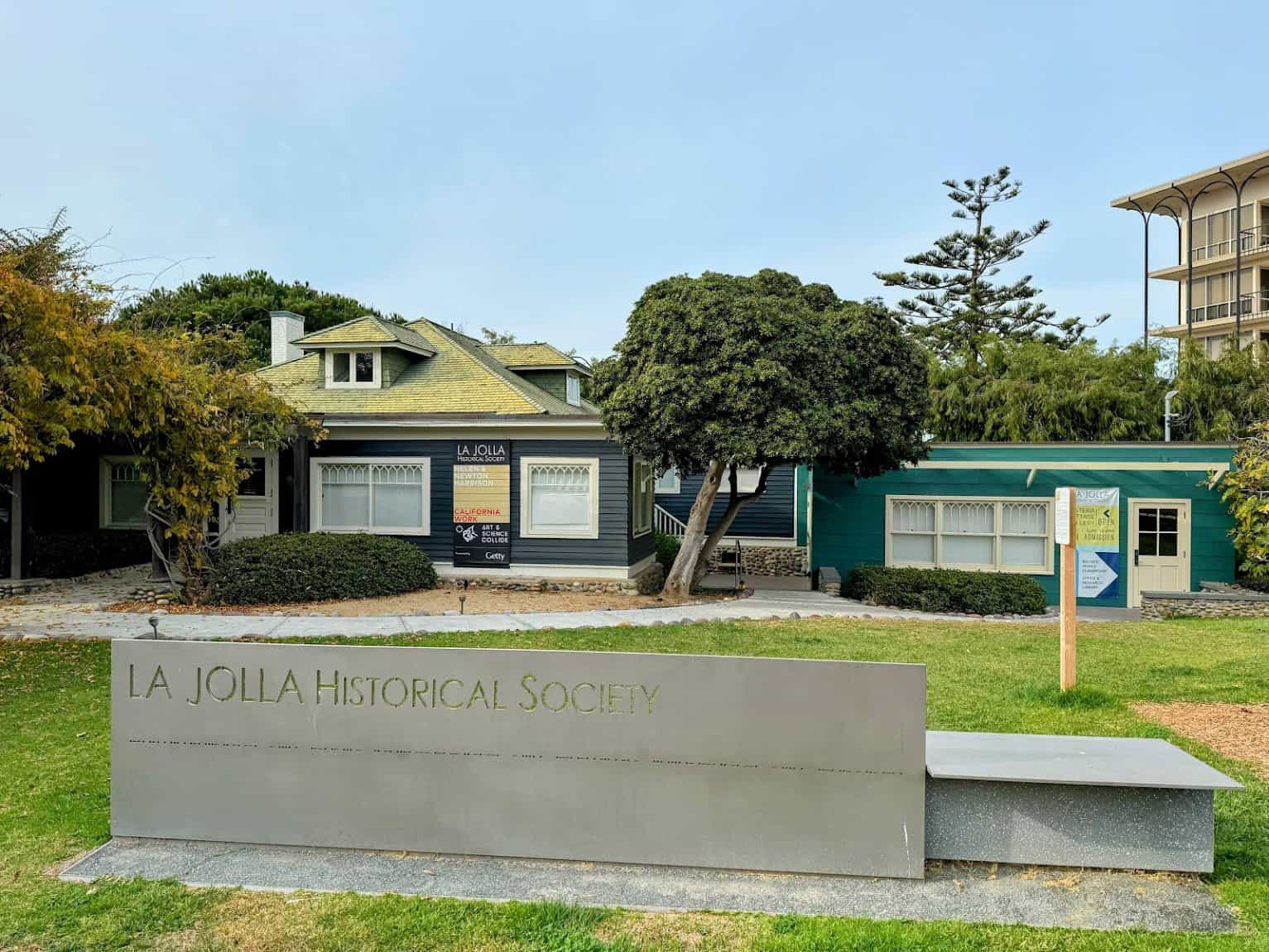 A historic house with charming architecture, featuring a green lawn and a modern sign that reads "La Jolla Historical Society." The building showcases classic window designs and a warm, inviting ambiance.