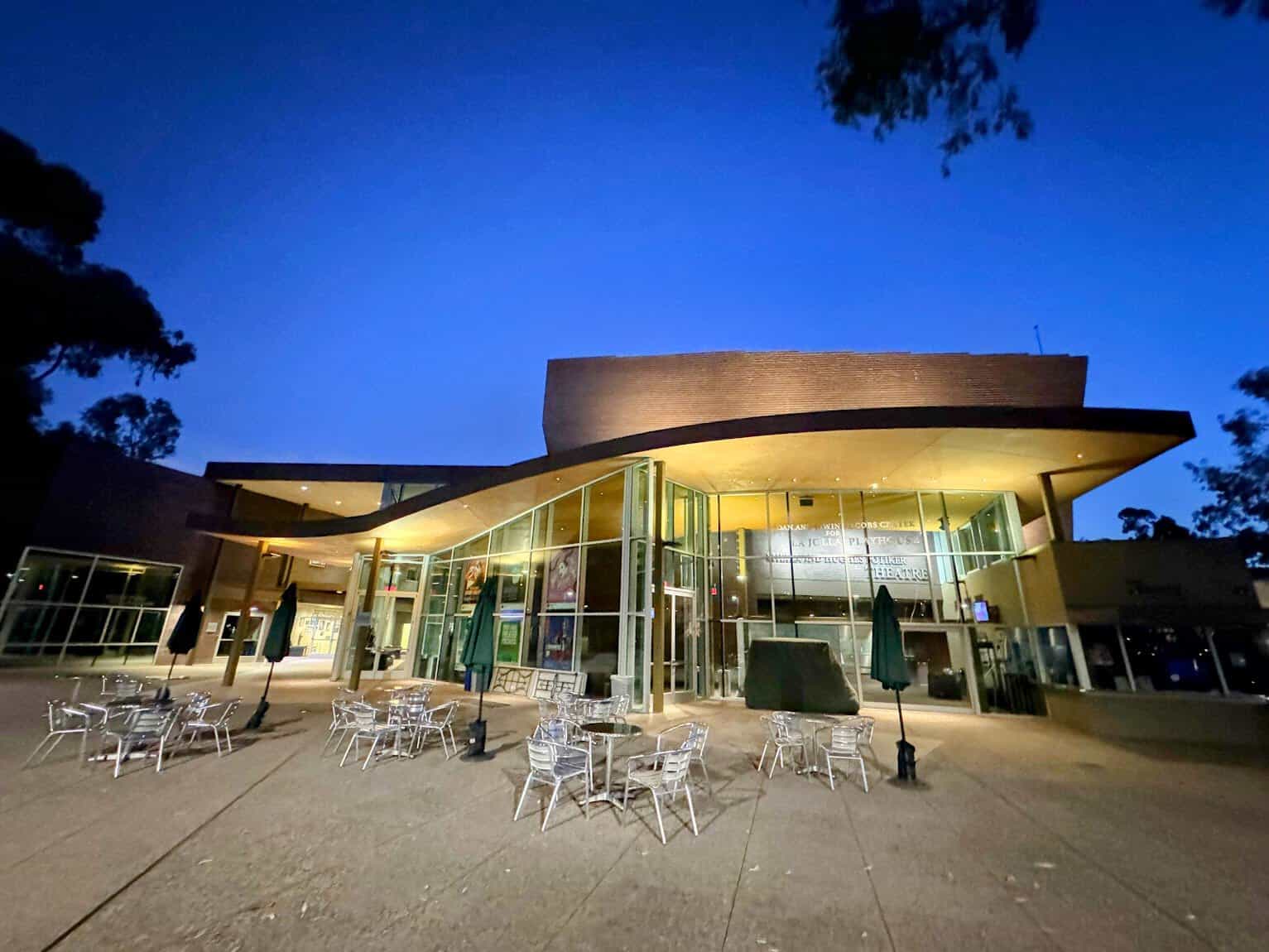 The modern exterior of the La Jolla Playhouse at twilight, with outdoor seating and glass-panel walls lit up to reveal its artistic ambiance.