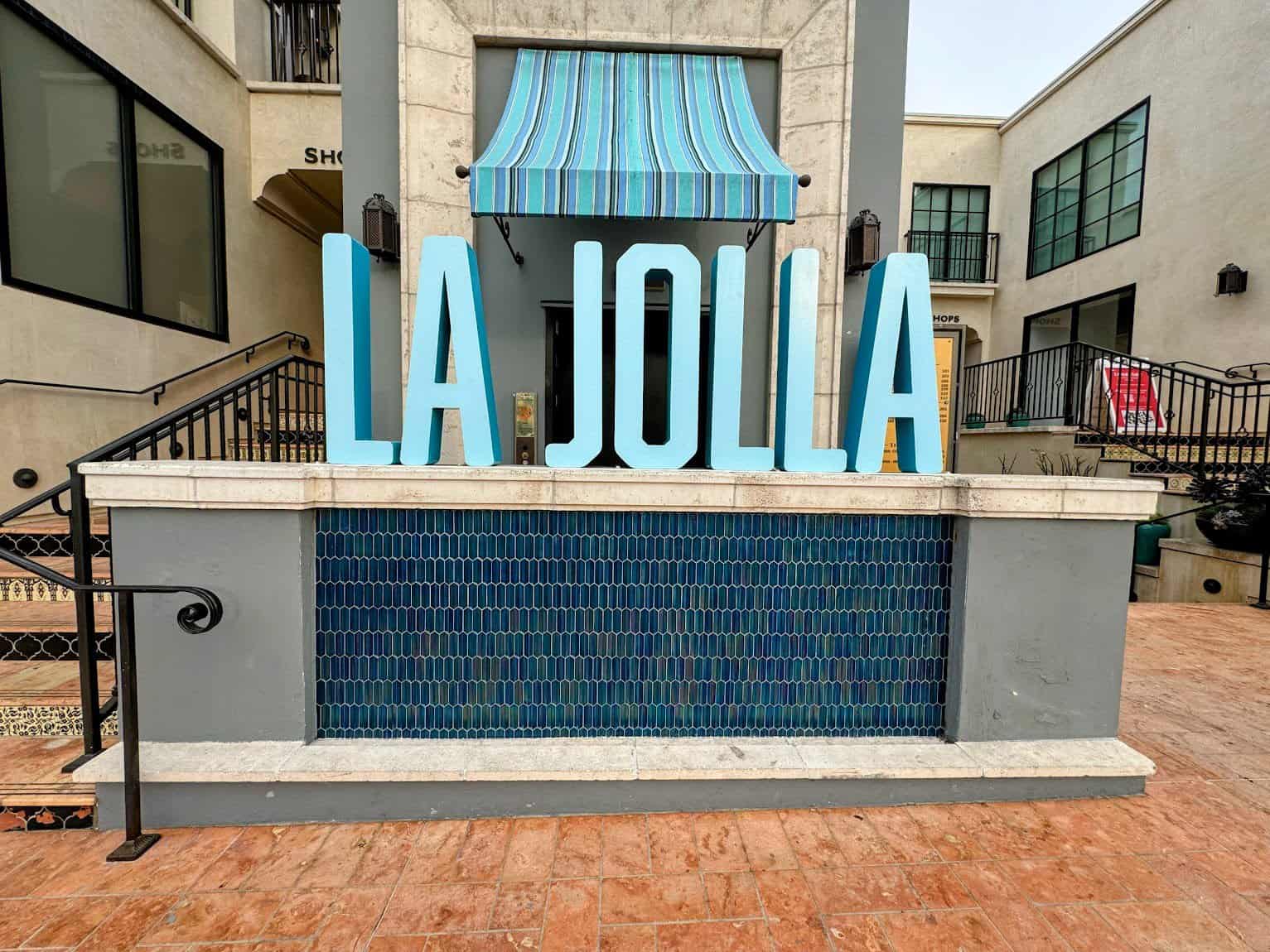 A bold and modern "La Jolla" sign in light blue letters, displayed on a tiled water feature in an outdoor shopping area.
