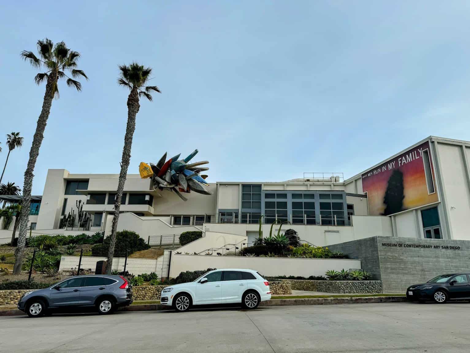 The modern facade of MCASD in La Jolla, featuring a unique sculpture of colorful surfboards and palm trees framing the building.