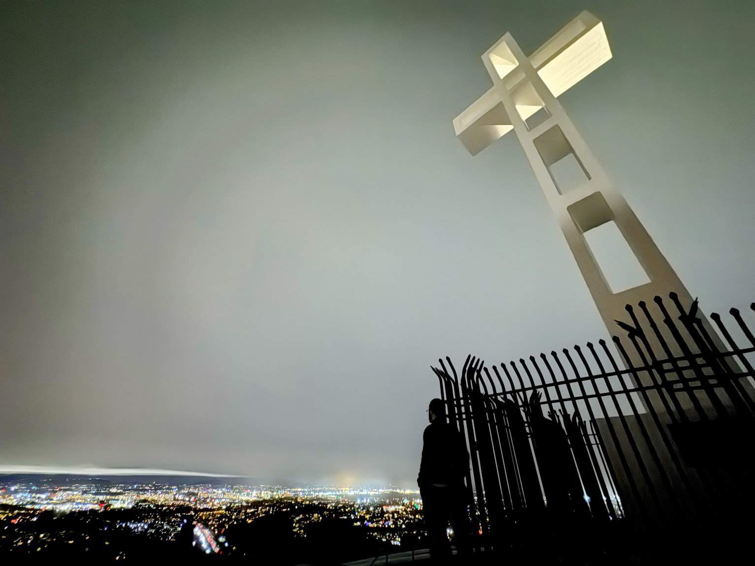 A large illuminated white cross on Mount Soledad against a hazy evening sky, with a view of city lights in the distance and silhouettes of visitors near the cross.