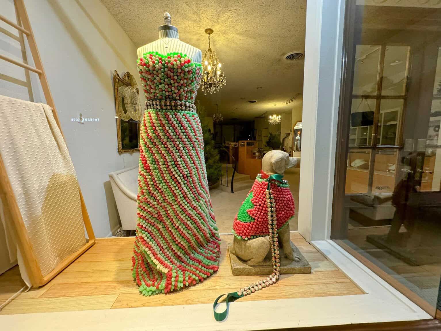 A festive shop window featuring a mannequin dressed in a colorful beaded gown and a dog sculpture wearing a matching outfit, creating a playful and elegant vibe.