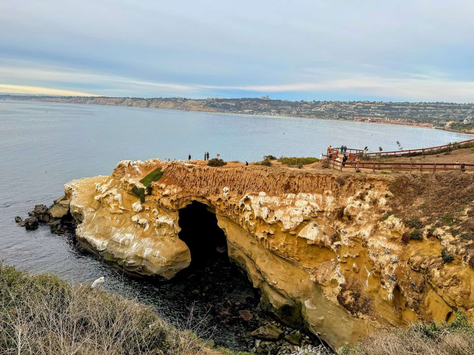 A large sandstone cave along the La Jolla coastline with an arched entrance opening to the ocean, surrounded by rugged cliffs and a walking path above.
