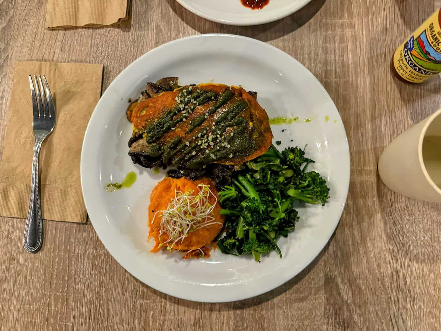 A healthy plant-based meal served on a white plate, featuring sweet potato mash topped with sprouts, sautéed broccoli, and a stuffed vegetable drizzled with green pesto sauce.