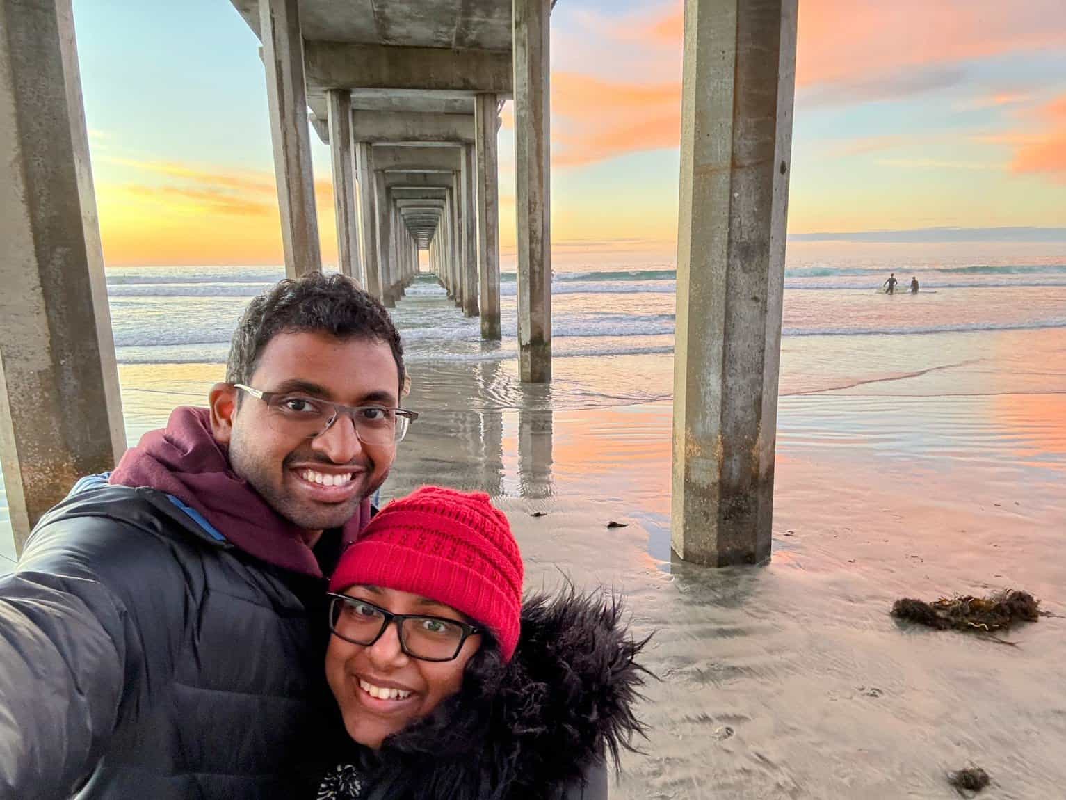 Kiran and Shreeyeh take a selfie under the UCSD Scripps Pier at sunset, with soft, colorful skies and ocean waves behind them.