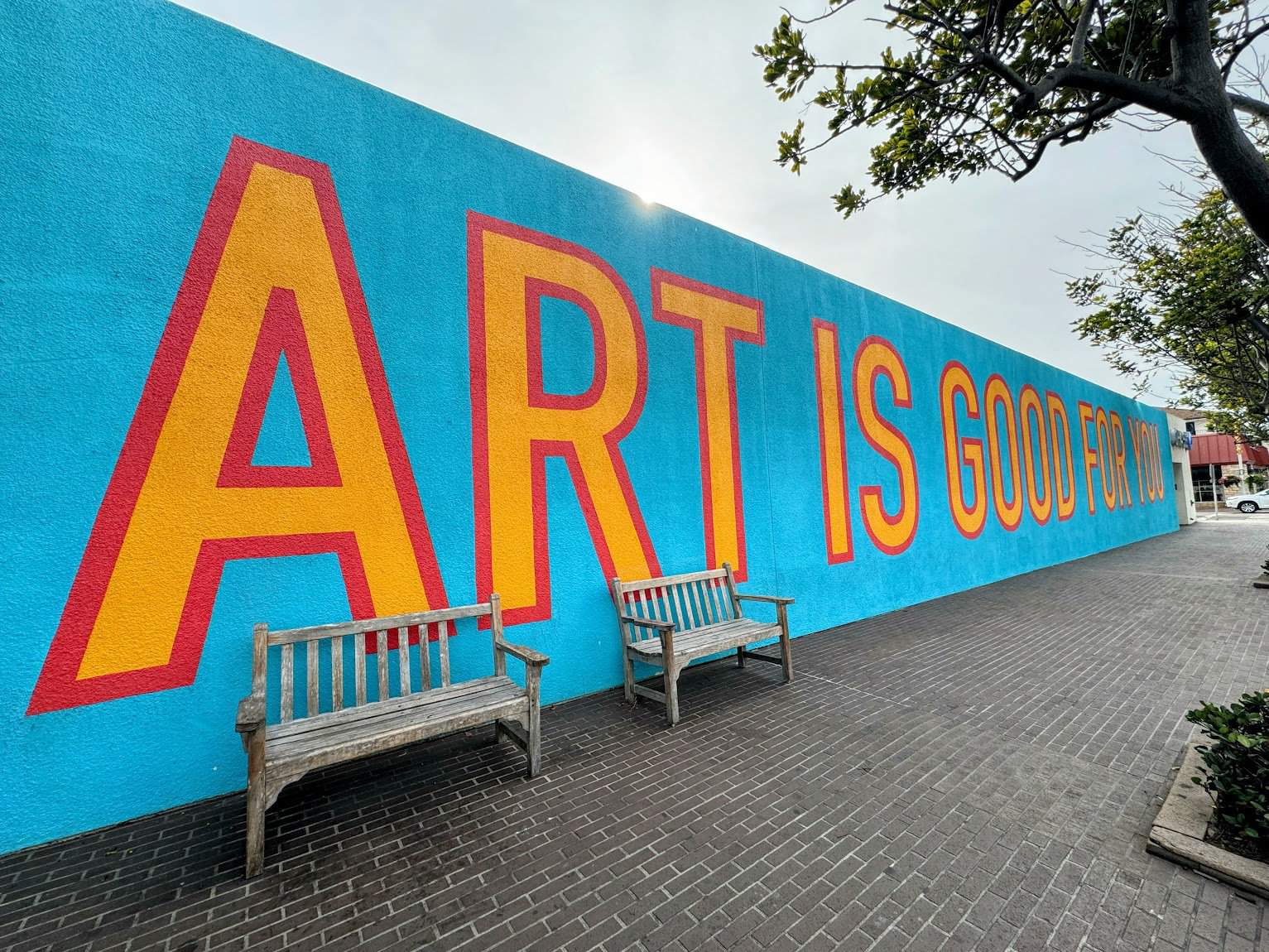 A bright blue wall with large text reading "ART IS GOOD FOR YOU" in bold yellow and red letters, accompanied by wooden benches on a brick walkway.