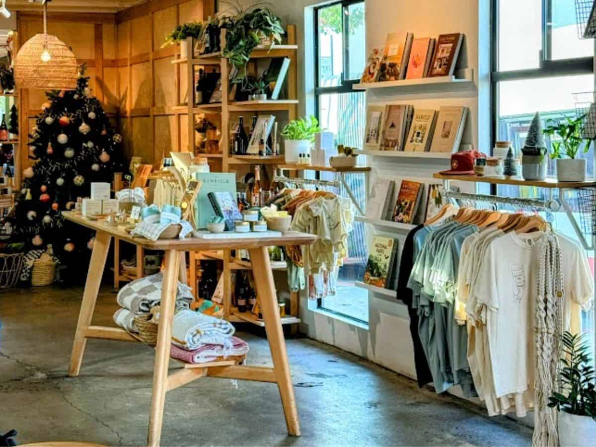 A trendy coffee shop with wooden shelving displays stocked with books, candles, home goods, and a small selection of T-shirts, with a cozy and curated vibe. A decorated Christmas tree glows softly in the background.