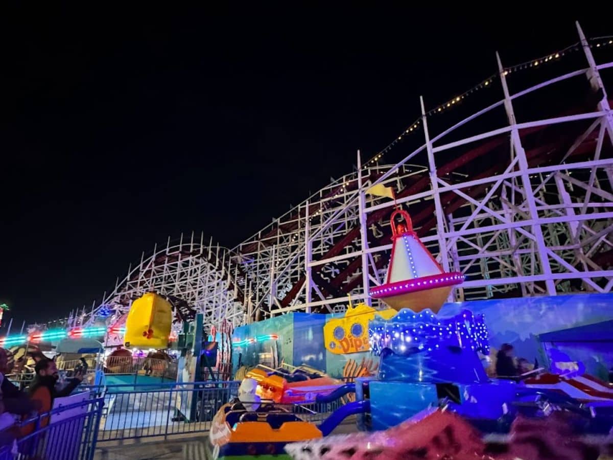A colorful nighttime scene at a San Diego amusement park with a classic wooden roller coaster in the background and vibrant kid-friendly rides in the foreground lit by string lights.
