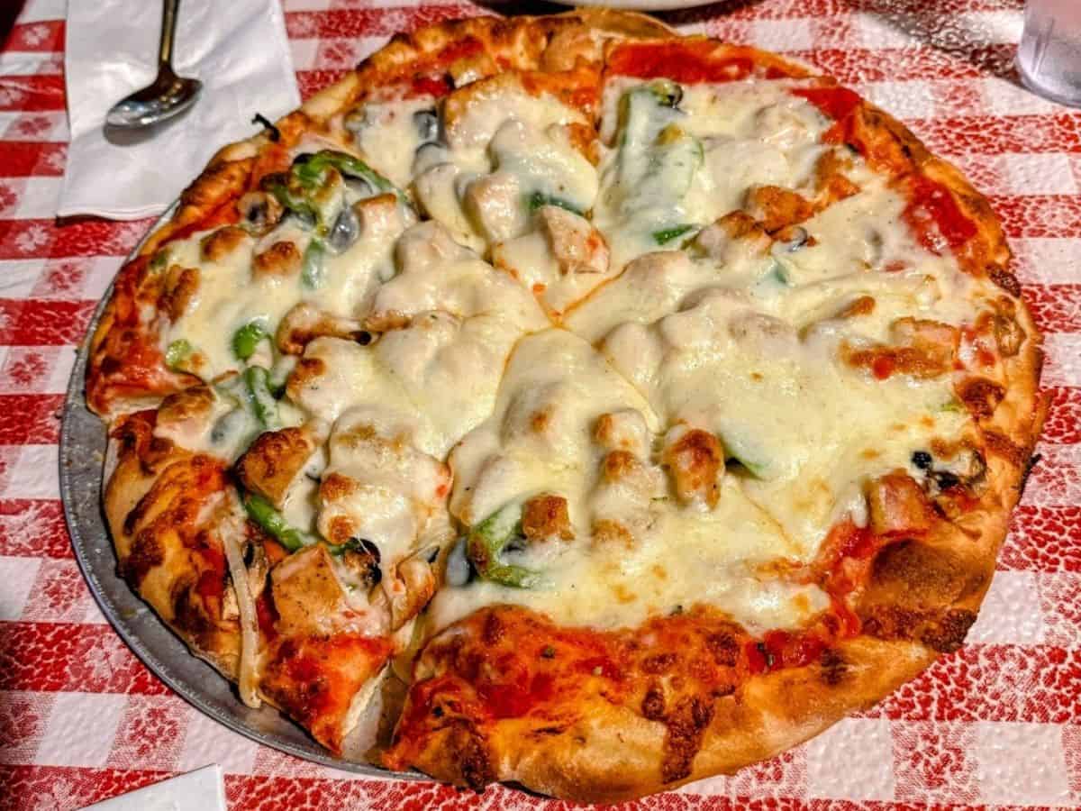 A classic, generously cheesed pizza loaded with green peppers, mushrooms, and chunks of grilled chicken sits on a metal tray over a red-and-white checkered tablecloth, giving off cozy, old-school pizzeria vibes.