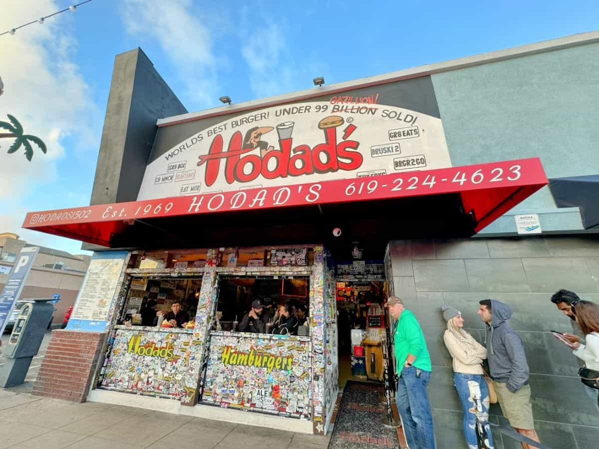 Front view of Hodad’s burger joint in San Diego with a long line of people waiting outside under a red awning that reads “World’s Best Burger! Under 99 Billion Sold!”