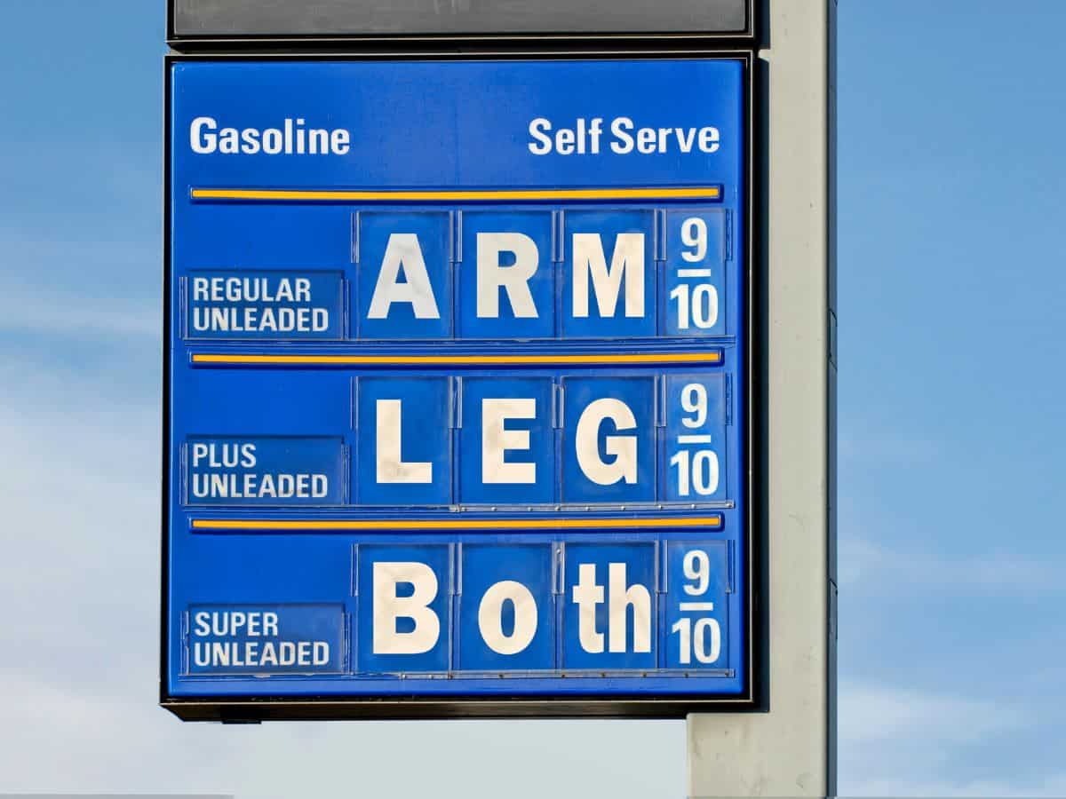 A blue gas station price sign displaying humorous labels instead of prices. The costs for "Regular Unleaded," "Plus Unleaded," and "Super Unleaded" are listed as "ARM," "LEG," and "Both," respectively, making a joke about high gas prices.