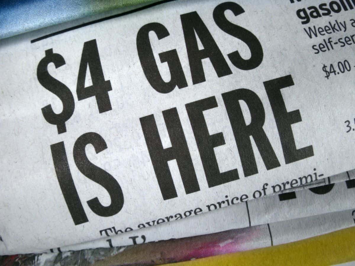 A close-up of a newspaper with a bold headline stating "$4 GAS IS HERE," emphasizing the rising cost of fuel. The text is printed in large black letters on a white background.