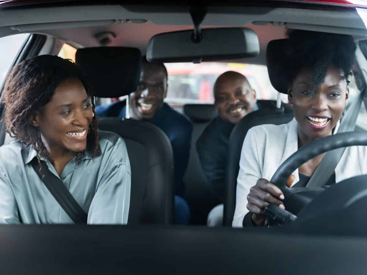 A group of four friends inside a car, laughing and chatting, with a woman in the driver’s seat smiling as she navigates.