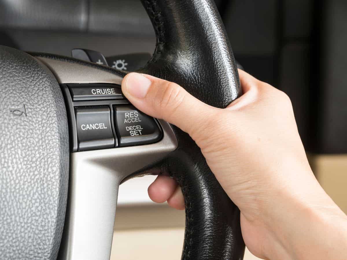 A hand pressing the cruise control button on a car’s steering wheel, labeled with "Cruise," "Cancel," "Res Accel," and "Decel Set."