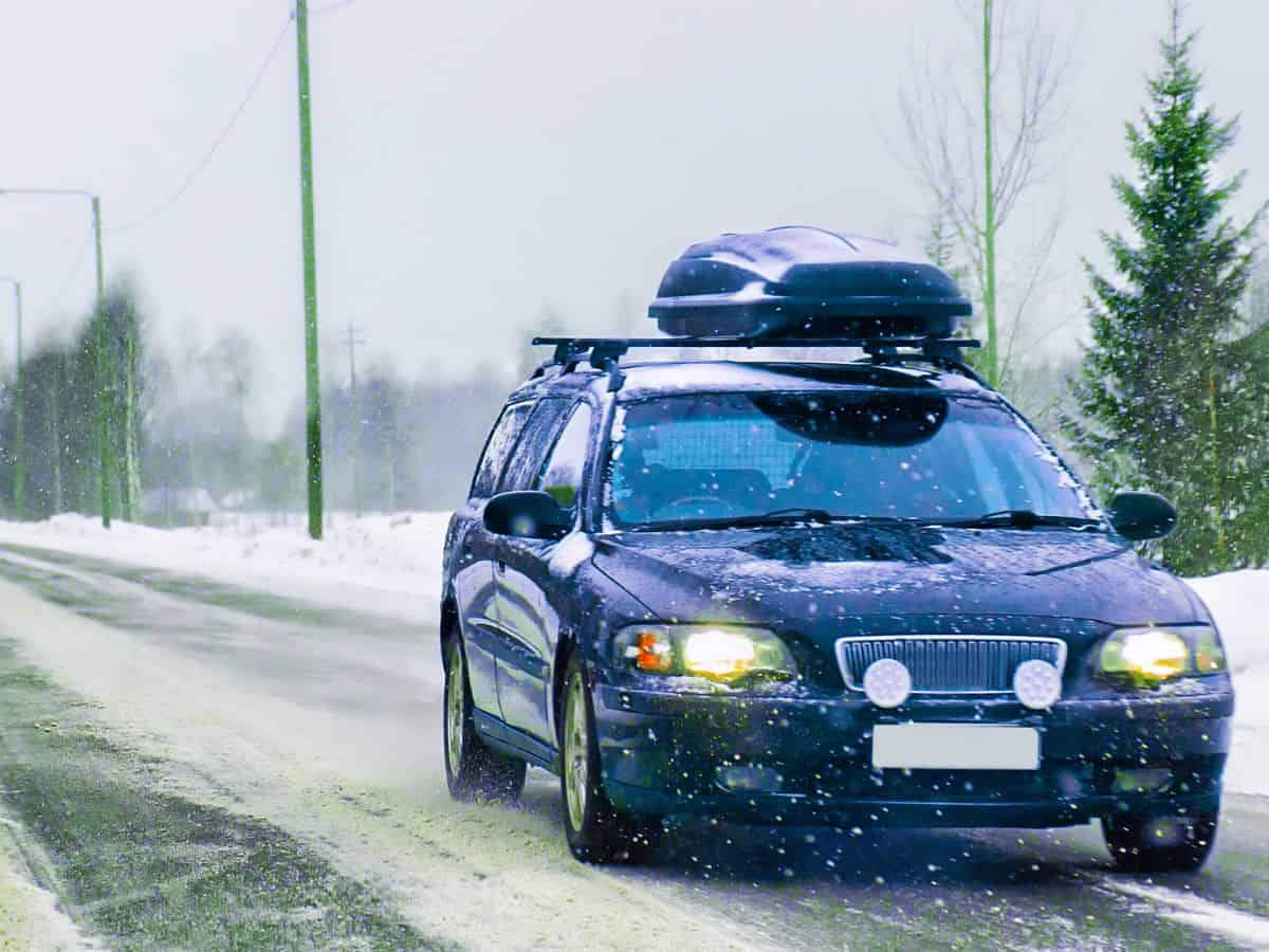 A black station wagon with a roof cargo box driving on a snowy road, headlights on, with snow-covered trees and power lines in the background.