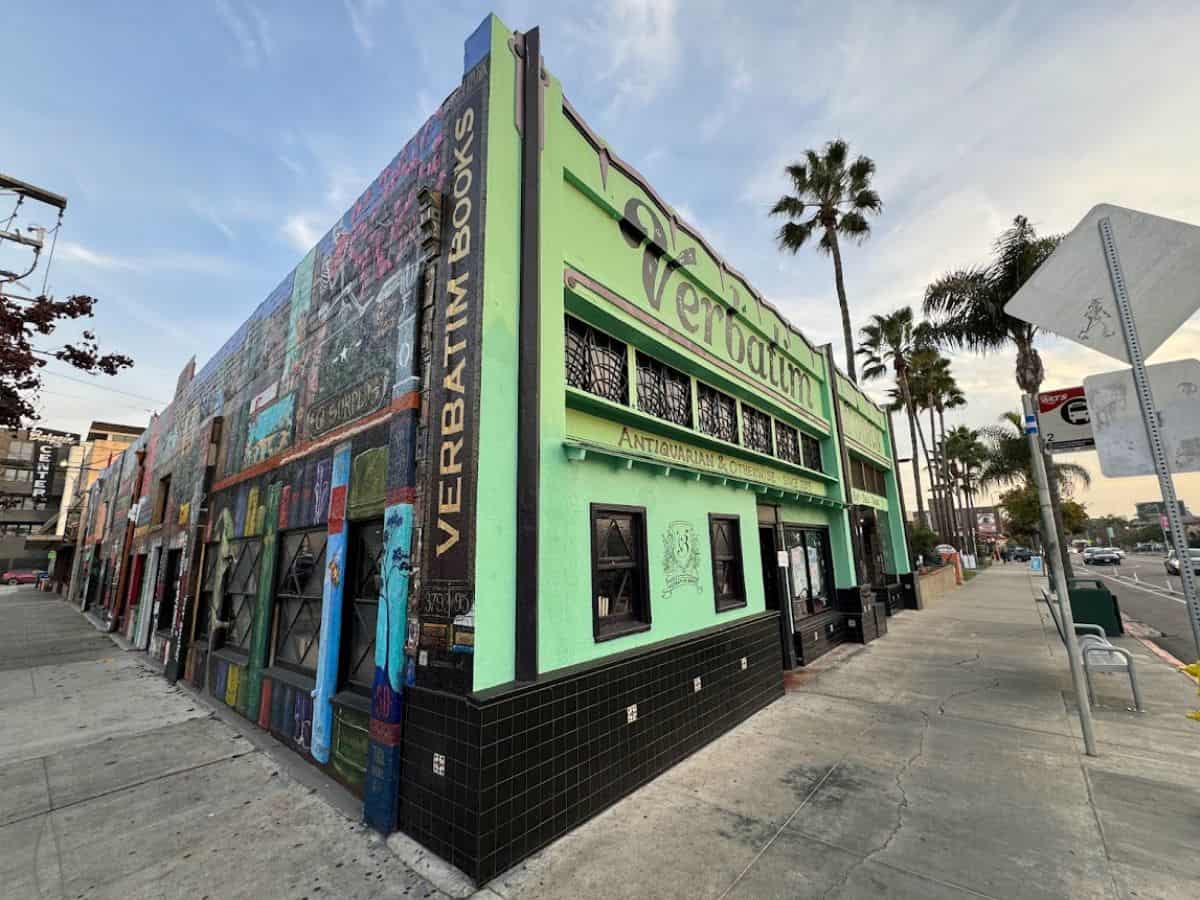 The exterior of Verbatim Books, an independent bookstore in San Diego, painted in bright green with a mural of books on its side. Palm trees line the street in the background.