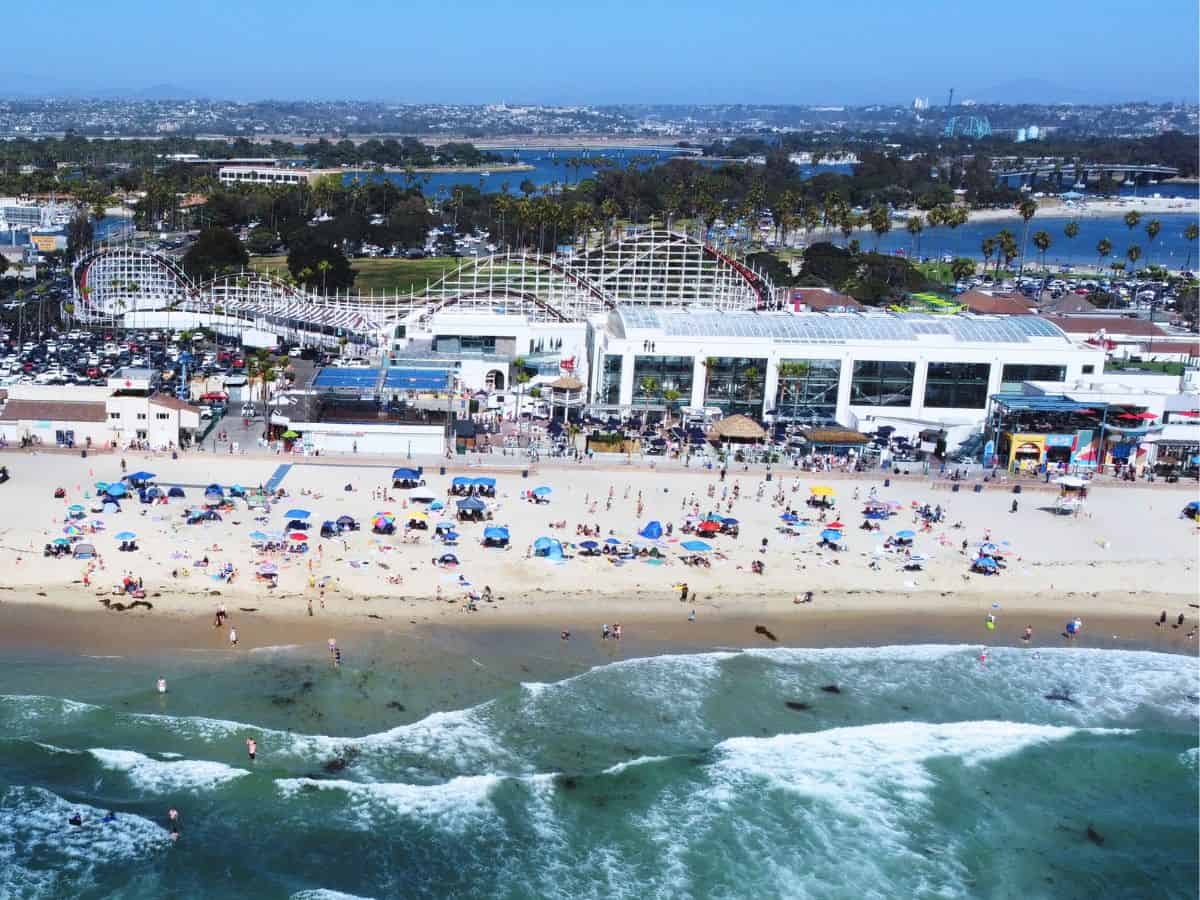 A sunny aerial view of a beachfront amusement park with a classic white wooden roller coaster and crowds on the sandy beach below. The ocean waves roll in while palm trees and parked cars fill the background.