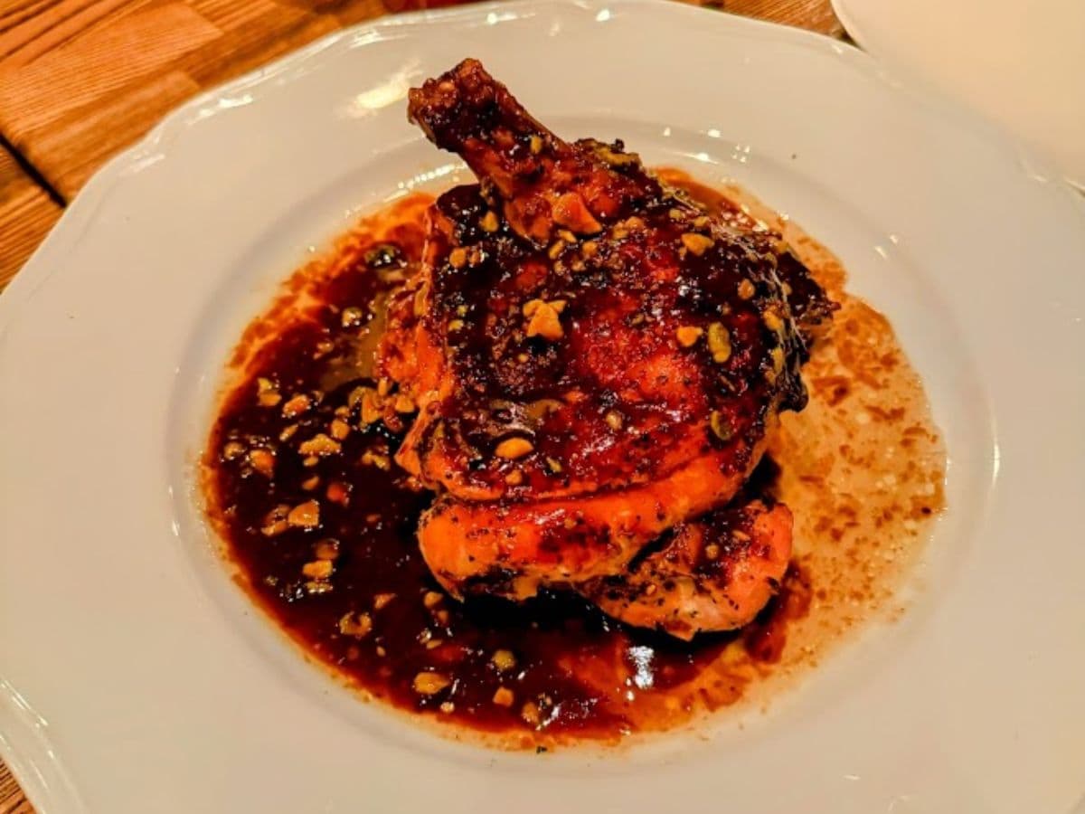 A close-up of a roasted chicken dish topped with a rich, dark sauce and chopped nuts, served on a white plate over a wooden table.