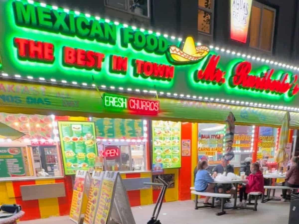 The colorful exterior of Mr. Ruribob's Mexican food restaurant lit up at night with bold neon signage reading "THE BEST IN TOWN," as diners eat at picnic tables outside under umbrellas.