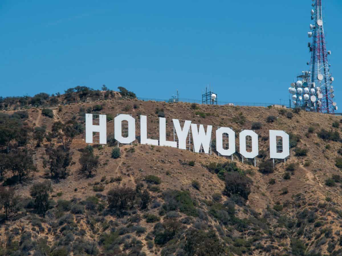 The iconic Hollywood Sign perched on the dry, rolling hills of Los Angeles, with a communications tower standing nearby. The white letters contrast sharply against the brown and green terrain.