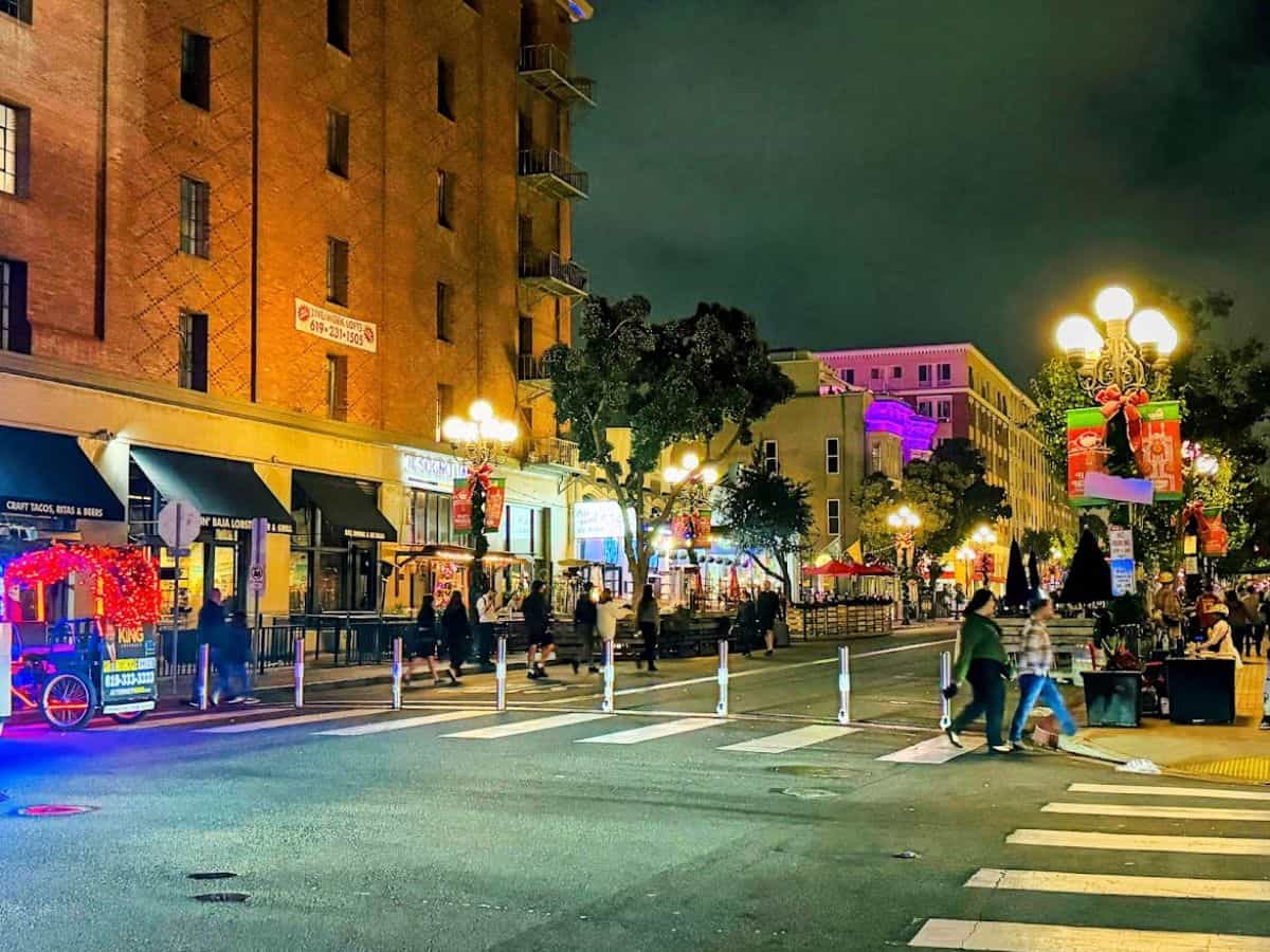 A lively San Diego street scene at night with holiday decorations, people walking, and restaurants and shops lit up in the Gaslamp Quarter.