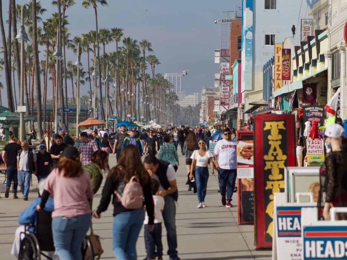 A lively Venice Beach Boardwalk scene with people walking past colorful storefronts and street vendors. Palm trees line the busy pathway under a sunny sky.
