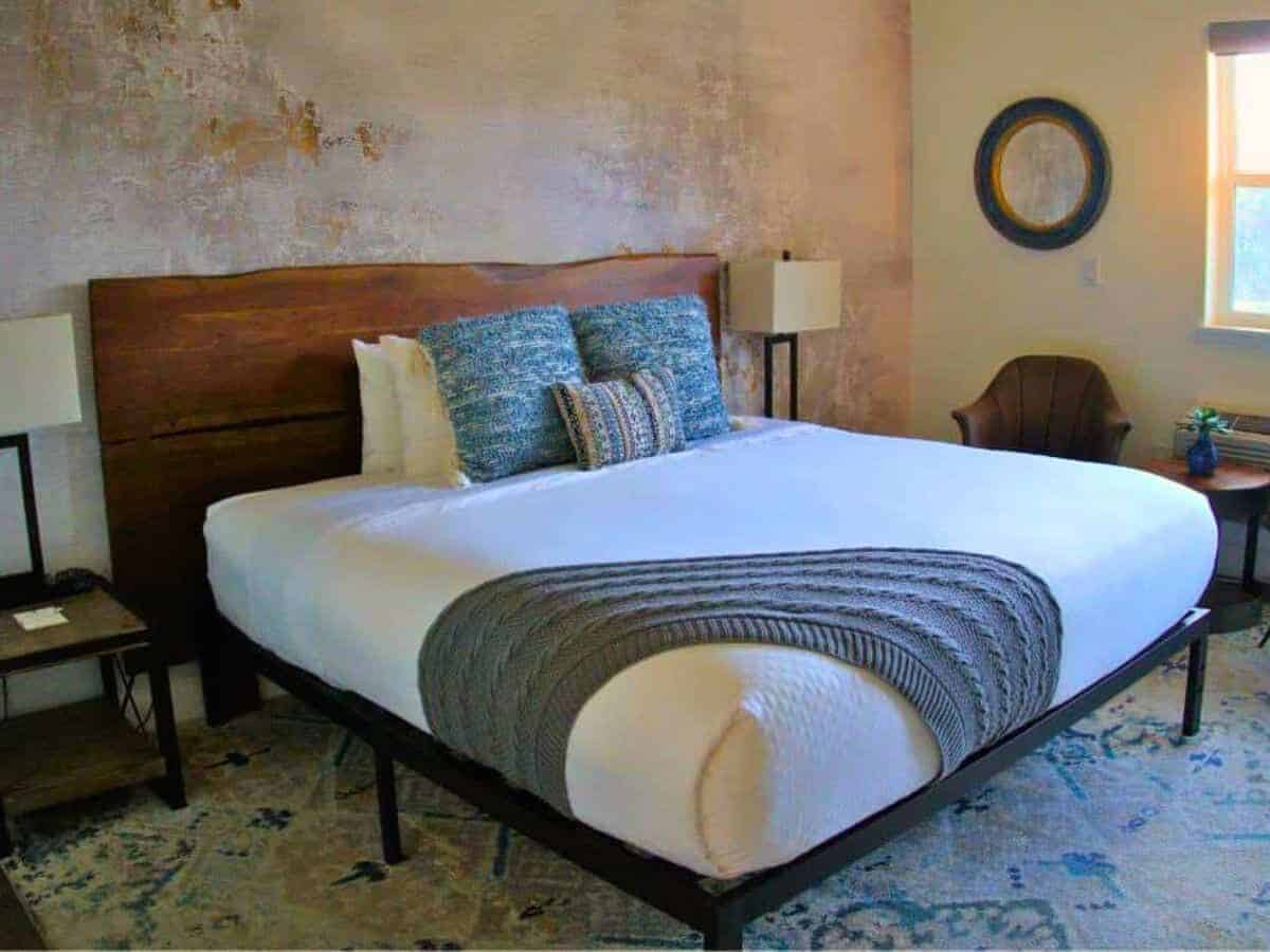 A cozy and stylish boutique hotel room with a wooden headboard, neatly made king-sized bed, textured accent wall, and warm ambient lighting.
