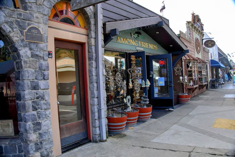 A charming stone-front boutique in Cambria, California, with seashell wind chimes and unique coastal gifts on display. The inviting storefront features a wooden sign and an "Open" sign on the door.