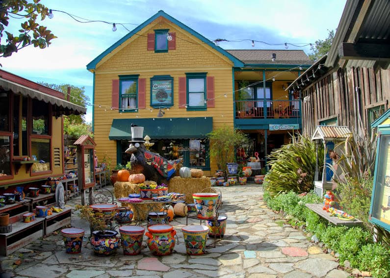 A vibrant, two-story yellow house with red shutters, surrounded by colorful pottery and festive autumn decorations. A bear statue in a top hat adds charm to the eclectic outdoor market.